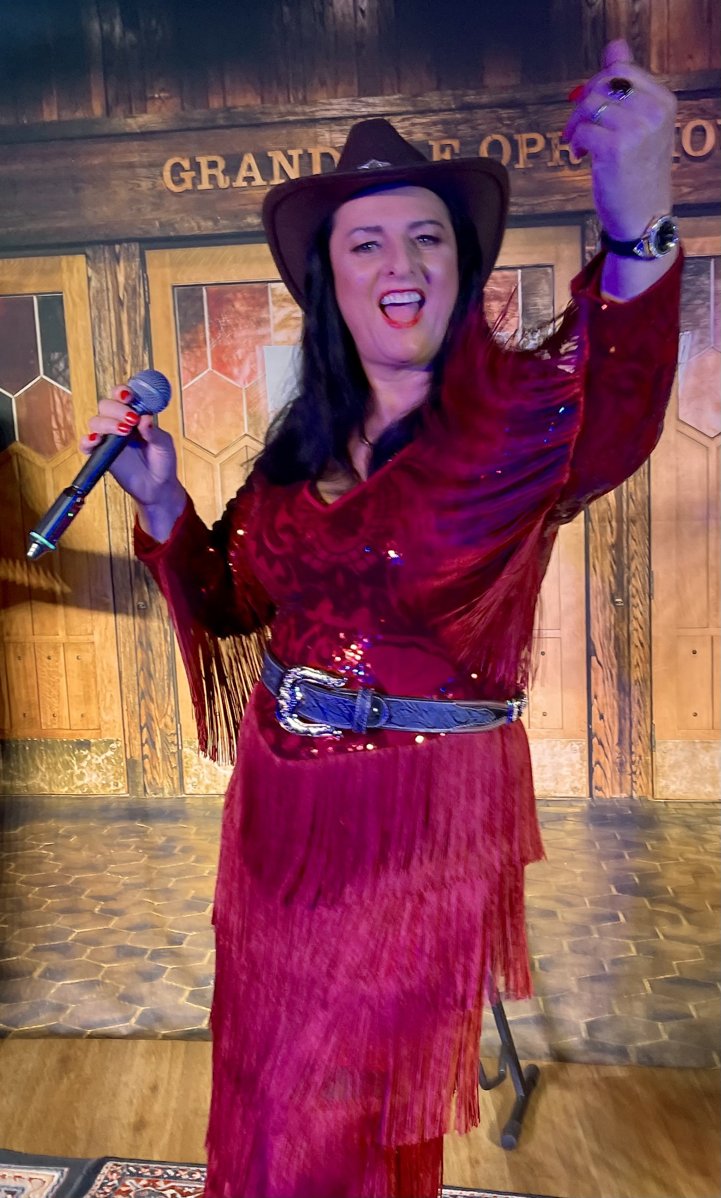 Jannine Douglas performing at the 2026 Country Set in a red sequin fringe dress and black cowboy hat at the Grand Ole Opry backdrop
