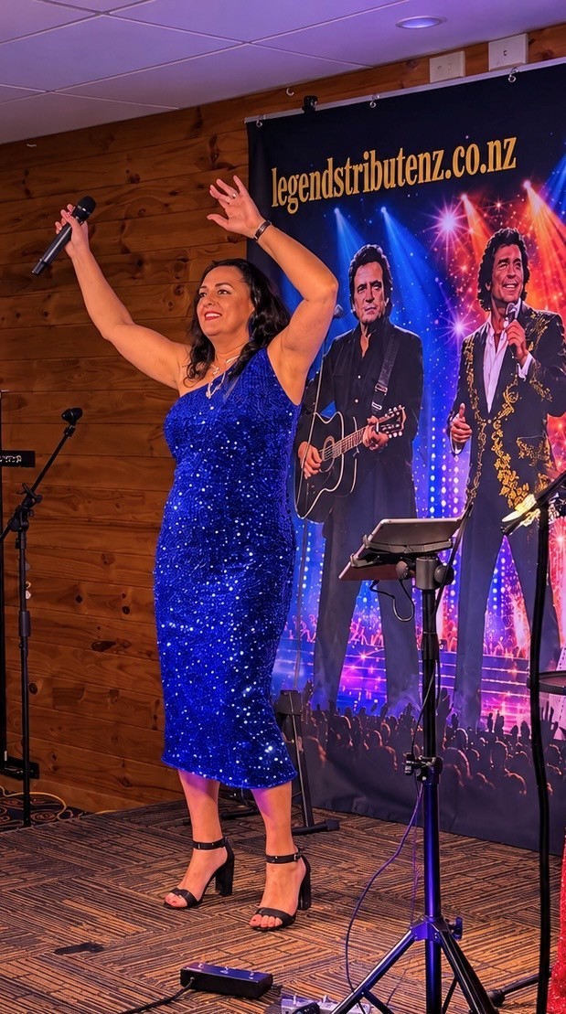 Jannine Douglas performing her dance set in a sparkling blue sequin dress at the Taupō show with the NZ Legends Tribute banner behind her
