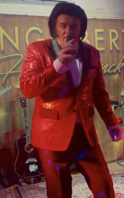 Trevor Needham performing as Engelbert Humperdinck in a red sequin suit with the Engelbert Humperdinck backdrop