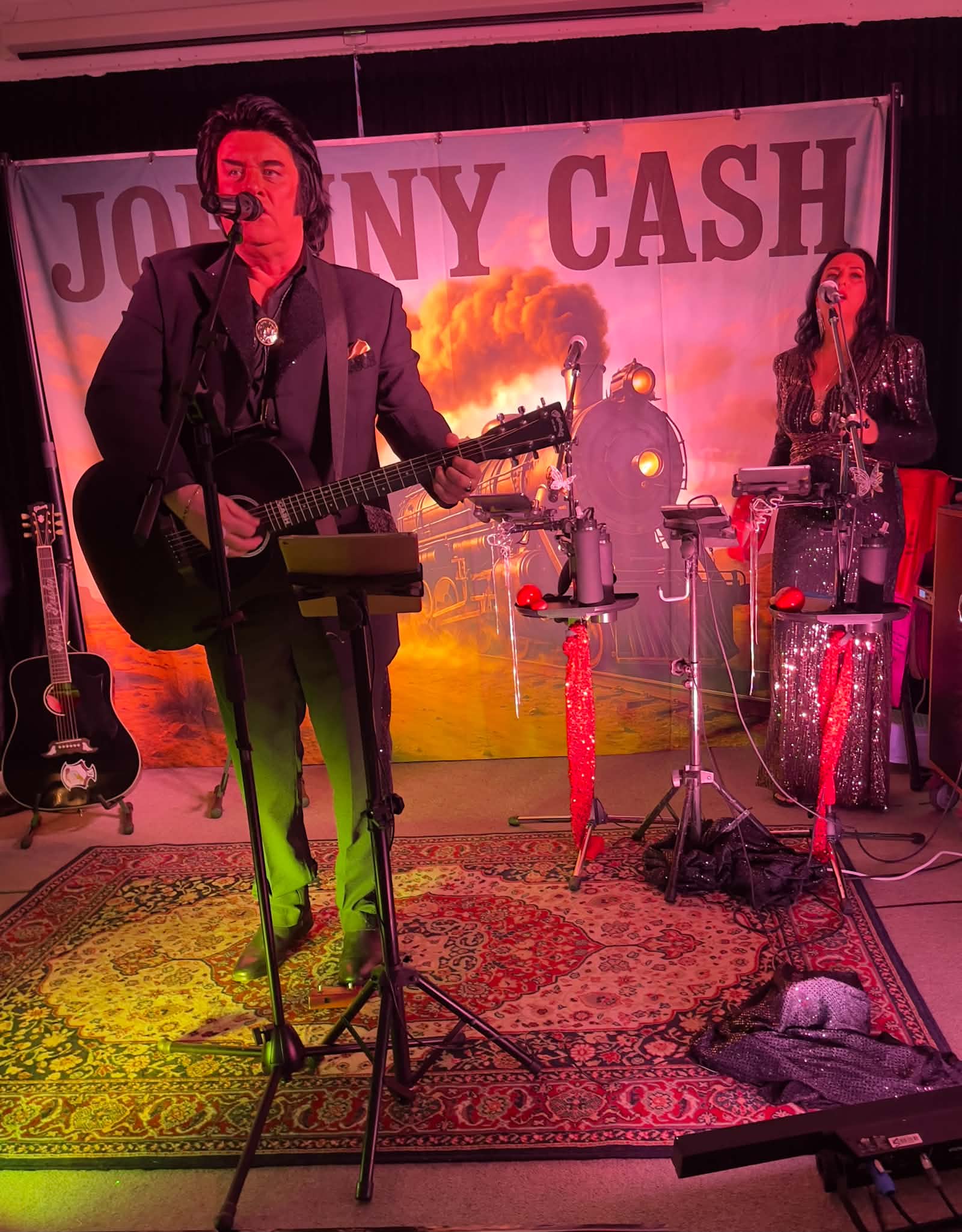 Trevor Needham as Johnny Cash with guitar and Jannine Douglas as June Carter performing live at Kamo 2026 with the Johnny Cash train backdrop