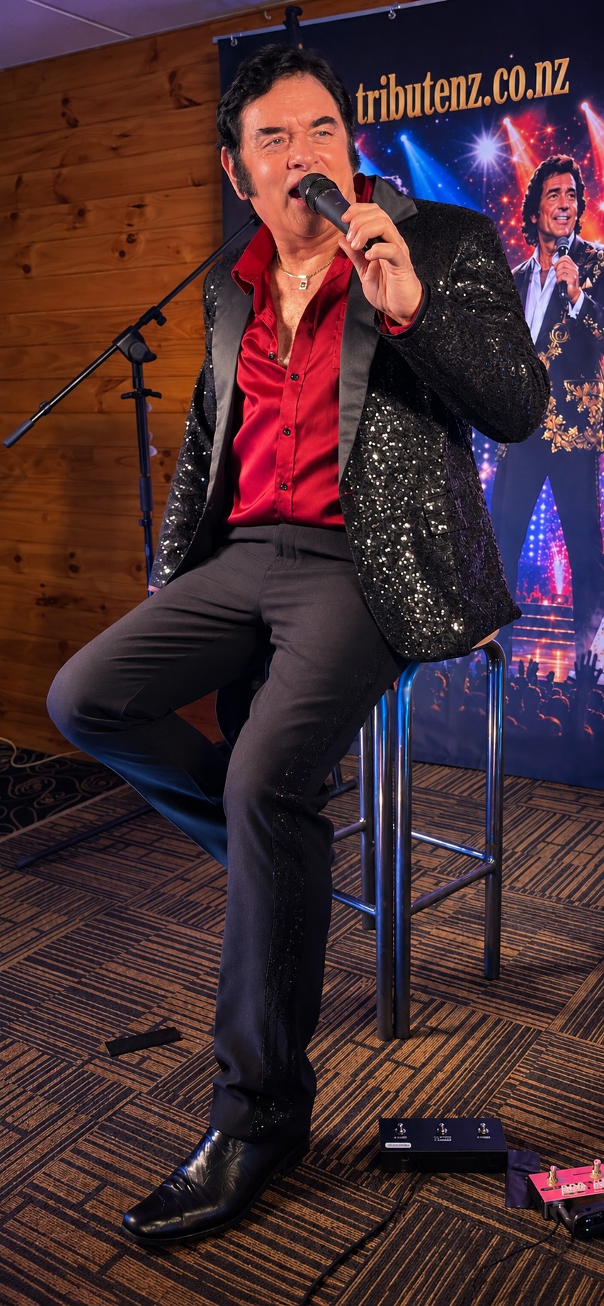 Trevor Needham as Neil Diamond seated on stage in a sparkle jacket and red shirt at Taupō Cosmopolitan Club